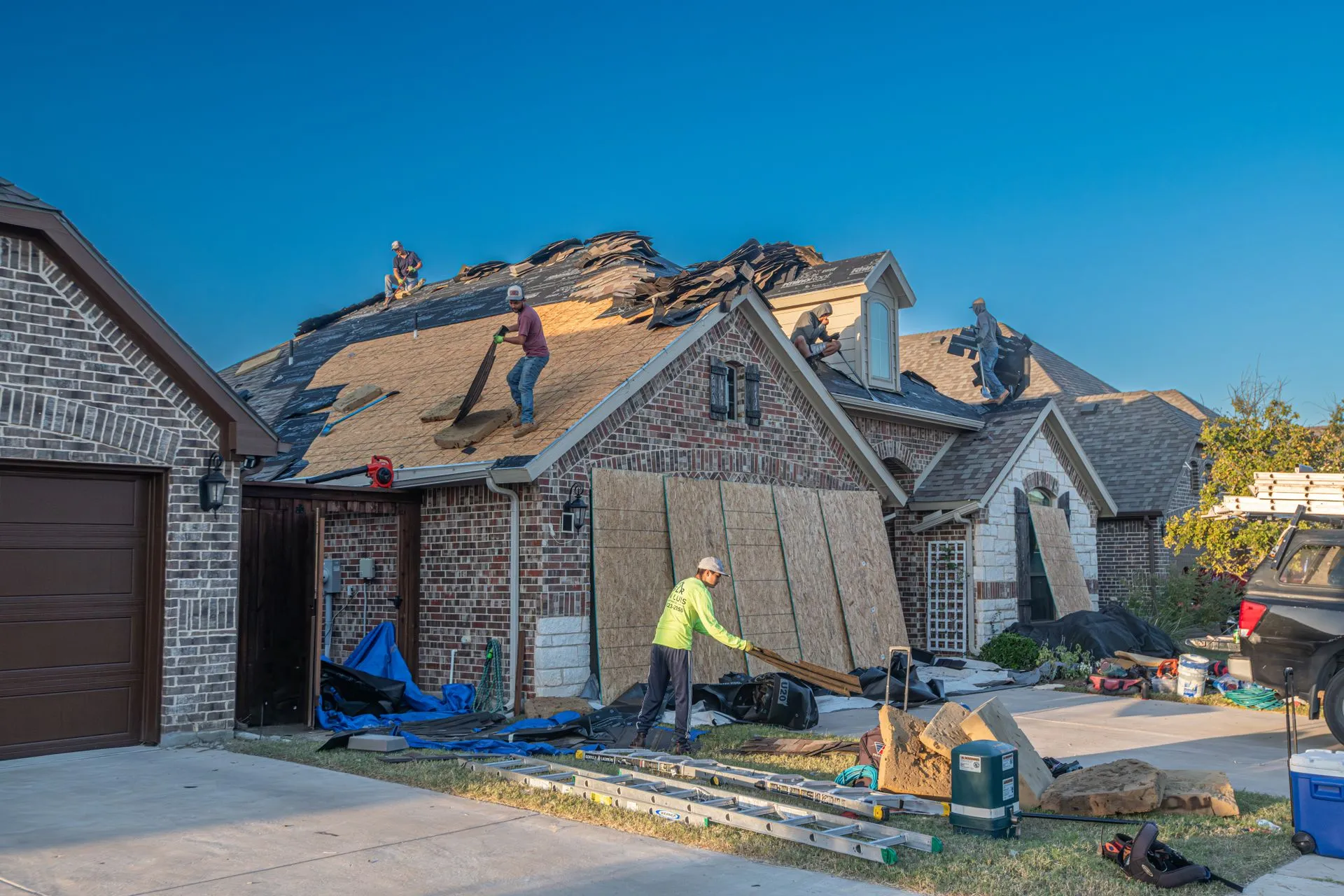 A roofing crew replacing the shingles on a brick house in a Fort Worth suburb, with the neighboring houses in view