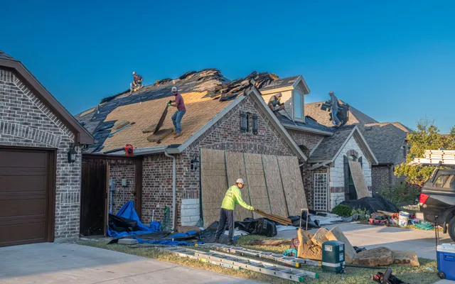 A roofing crew replacing the shingles on a brick house in a Fort Worth suburb, with the neighboring houses in view